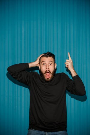 Portrait of young man with shocked facial expression pointing up over blue studio backgroundの写真素材