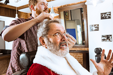 The senior man in Santa claus costume shaving his personal master at barber shop before Christmasの写真素材