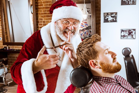 The senior man in Santa claus costume working as personal master with scissors at barber shop before Christmasの写真素材