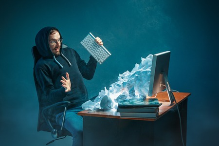 Young stressed handsome businessman working at desk in modern office shouting at laptop screen and...