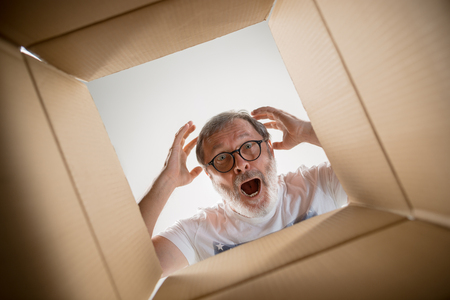 The surprised senior man unpacking, opening carton box and looking inside. The package, delivery, surprise, gift lifestyle concept. Human emotions and facial expressions conceptsの写真素材