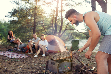 Party, camping of men and women group at forest. They relaxing, singing a song and cooking barbecue against green grass. The vacation, summer, adventure, lifestyle, picnic conceptの写真素材
