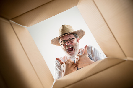 The surprised senior man unpacking, opening carton box and looking inside. The package, delivery, surprise, gift lifestyle concept. Human emotions and facial expressions conceptsの写真素材