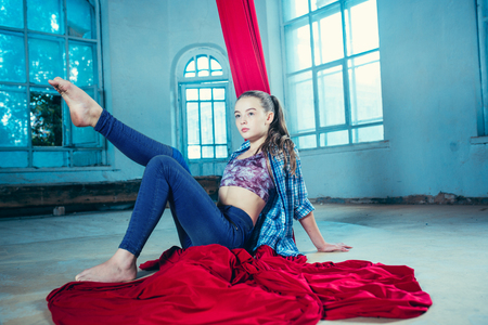 Graceful gymnast resting after performing aerial exercise with red fabrics on blue old loft background. Young teen caucasian fit girl. The circus, acrobatic, acrobat, performer, sport, fitness, gymnastic conceptの写真素材