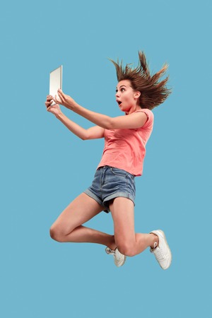 Jump of young woman over blue studio background using laptop or tablet gadget while jumping. Runnin girl in motion or movement. Human emotions and facial expressions concept. Gadget in modern lifeの写真素材