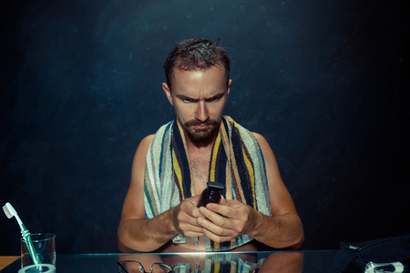 The young caucasian man in bedroom sitting in front of the mirror during scratching his beard at home. Human emotions concept. Lifestyle and skincare conceptsの写真素材