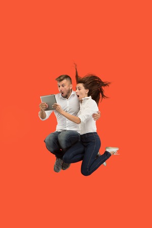 Jump of young couple over red studio background using laptop or tablet gadget while jumping. Runnin...