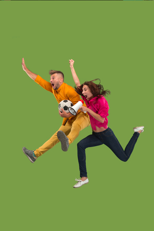 Forward to the victory.The young couple as soccer football player jumping and kicking the ball at studio on a green background. Football fan and world championship concept. Human emotions conceptsの写真素材