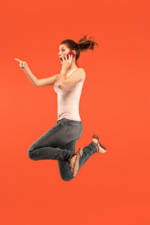 always on mobile. Full length of pretty young woman taking phone while jumping against red studio background. Mobile, motion, movement, business conceptsの写真素材