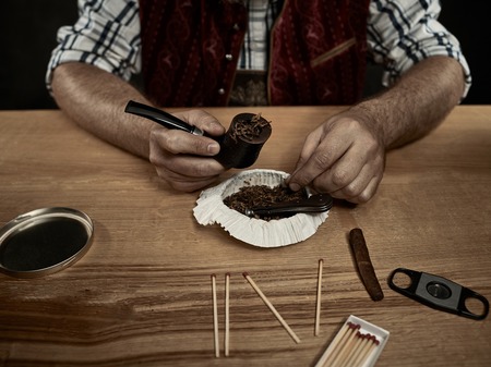bearded man clogs the tobacco in pipe. The male hands close upの写真素材