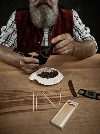 The senior bearded man sitting at table and clogging the tobacco in pipe. The male hands close up. Bavaria. a man dressed in traditional Bavarian or Austrian national traditional costumeの写真素材