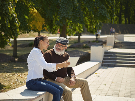 Help someone you love. Portrait of young smiling girl sitting with grandfather and laptop against city park. community and family lifestyle conceptの写真素材