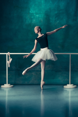 The classic ballet dancer in white tutu posing at ballet barre on studio background. Young teen before dancing. Ballerina project with caucasian model. The ballet, dance, art, contemporary, choreography conceptの写真素材