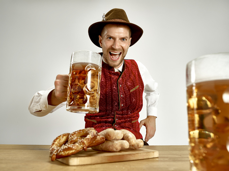 Germany, Bavaria, Upper Bavaria. The young happy smiling man with beer dressed in traditional Austrian or Bavarian costume holding mug of beer at pub or studio. The celebration, oktoberfest, festival conceptの素材 [FY310107876061]