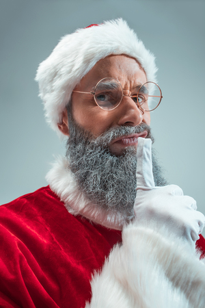 Funny serious guy with christmas hat posing at studio. New Year Holiday. Christmas, x-mas, winter, gifts concept. Man wearing Santa Claus costume on gray. Copy space. Winter sales.の写真素材
