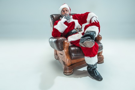 Funny serious guy with christmas hat sitting at studio. New Year Holiday. Christmas, x-mas, winter, gifts concept. Man wearing Santa Claus costume on gray. Copy space. Winter sales.の写真素材