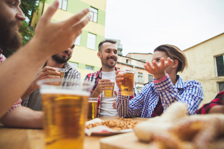 The group of friends enjoying and drinking beer at outdoor bar or pub. Boiled white sausages, served with beer and pretzels. Perfect for Octoberfest. Natural wooden background. Front view with female and male handsの写真素材