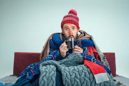 Bearded sick man with flue sitting on sofa at home or studio with cup of tea covered with knitted warm clothes. Illness, influenza concept. Relaxation at Home. Healthcare Concepts.の写真素材