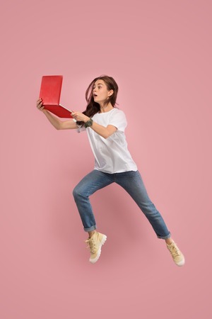 Gadget in modern life. Jump of young woman over pink studio background using laptop or tablet gadget while jumping. Runnin surprised girl in motion or movement. Human emotions and facial expressions concept.の写真素材