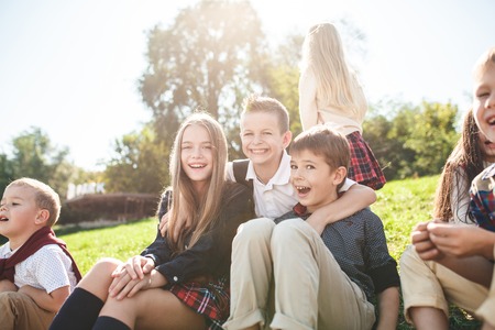 A group of happy smiling children of school and preschool age are sitting on the green grass in the...