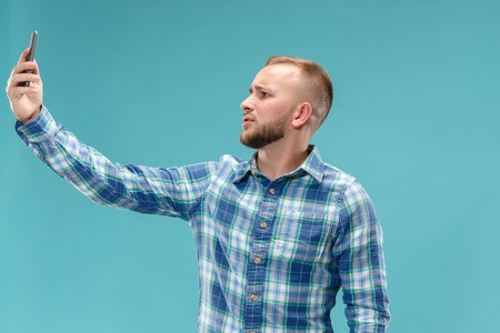 Portrait of attractive serious young man taking a selfie with his smartphone. Isolated on blue studio background. Human emotions, facial expression concept. Trendy colorsの写真素材