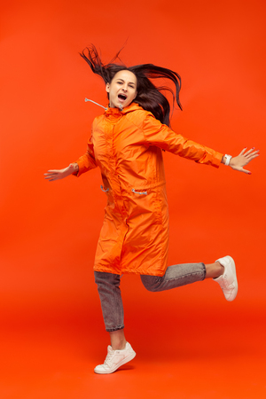 The happy smiling young girl posing at studio in autumn orange jacket isolated on red.の写真素材