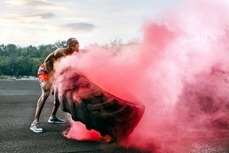 Handsome african american muscular man flipping burning big tire outdoor with smokeの写真素材