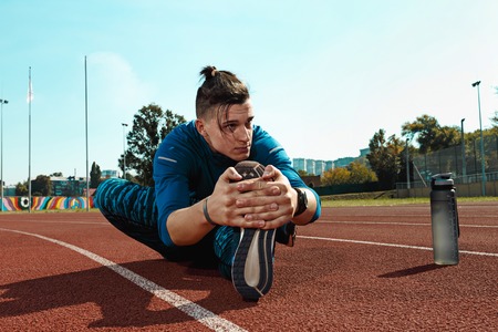 The fit male runner stretching legs preparing for run during training at stadium tracks. The athlete, fitness, workout, sport, exercise, training, athletic, lifestyle conceptの写真素材