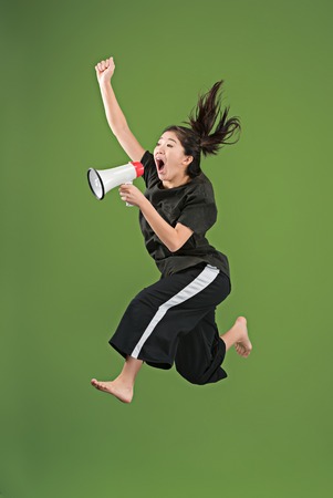 Beautiful young woman jumping with megaphone isolated over green background. Runnin girl in motion or movement. Human emotions and facial expressions conceptの写真素材
