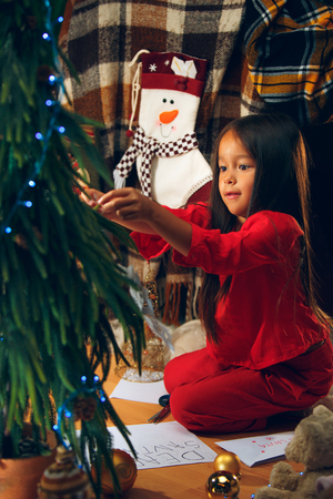 christmas, holidays and childhood concept - happy girl in red clothes decorating natural fir tree at homeの写真素材