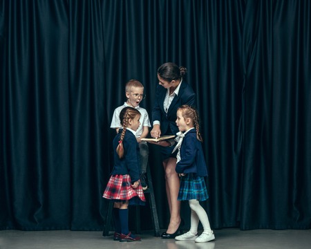 Cute smiling happy stylish children and female teacher on dark background. Beautiful stylish teen girls and boy standing together and posing on the school stage in front of the curtain. Classic style. Kids fashion and emotions concept.の写真素材