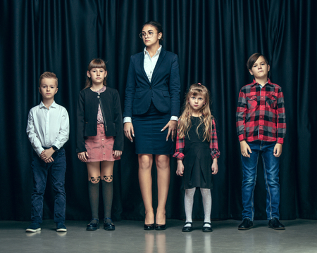 Cute smiling happy stylish children and female teacher on dark background. Beautiful stylish teen girls and boy standing together and posing on the school stage in front of the curtain. Classic style. Kids fashion and emotions concept.の写真素材