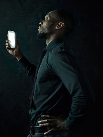 Studio shot of young serious black African man thinking while talking on mobile phone against black studio backgroundの写真素材