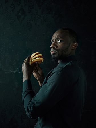 The young african american man eating hamburger and looking away on black studio background