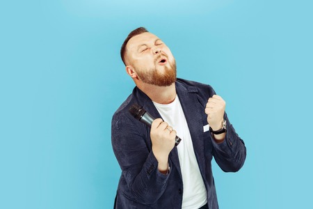 Young man with microphone on blue studio background, leading concept. Human emotions and facial...