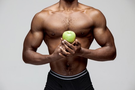 Fit young man with beautiful torso with green apple isolated on gray background. The naked torso of African American man posing at studio. The body, fitness, sports, healthy lifestyle and bodybuilder concept.の写真素材