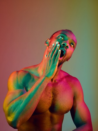 Close up portrait of a young naked yawning african man. High Fashion male model in colorful bright lights posing in studio. Art design over vivid background.の写真素材