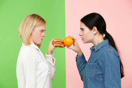 Diet. Dieting concept. Healthy useful food. Beautiful young women choosing between fruits and unhelathy fast food at studio. Human emotions and comparison conceptsの写真素材