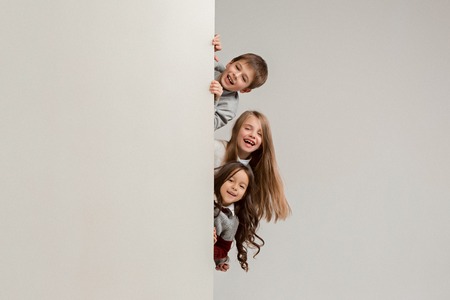 Banner with a surprised children peeking at the edge with copyspace. The portrait of cute little kids boy and girls looking at camera against white studio wall. Kids fashion and happy emotions conceptの写真素材