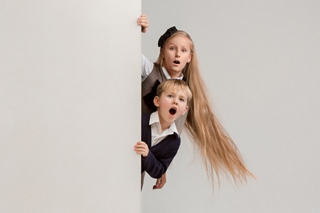Banner with a surprised children peeking at the edge with copyspace. The portrait of cute little kids boy and girls looking at camera against white studio wall. Kids fashion and happy emotions conceptの写真素材