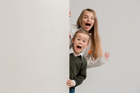 Banner with a surprised children peeking at the edge with copyspace. The portrait of cute little kids boy and girls looking at camera against white studio wall. Kids fashion and happy emotions conceptの写真素材