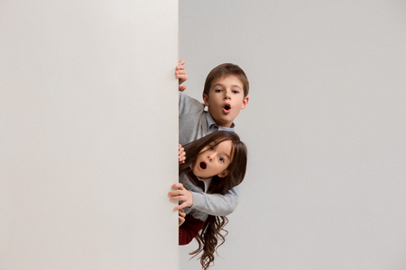 Banner with a surprised children peeking at the edge with copyspace. The portrait of cute little kids boy and girls looking at camera against white studio wall. Kids fashion and happy emotions conceptの写真素材