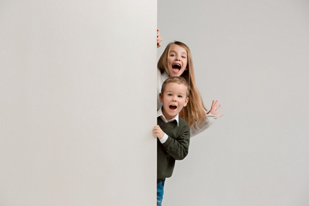 Banner with a surprised children peeking at the edge with copyspace. The portrait of cute little kids boy and girls looking at camera against white studio wall. Kids fashion and happy emotions conceptの写真素材
