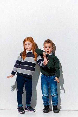 The portrait of cute little kids boy and girl in stylish jeans clothes looking at camera against white studio wall. Kids fashion and happy emotions conceptの写真素材