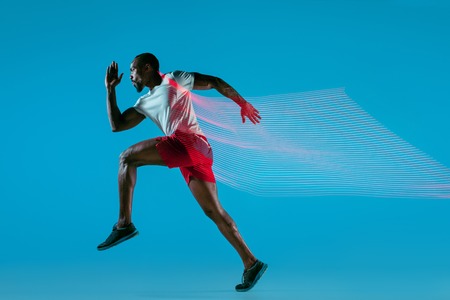 Full length portrait of active young african muscular running man, isolated over blue studio background with flashes of lightの写真素材