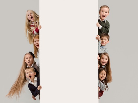 Banner with a surprised children peeking at the edge with copyspace. The portrait of cute little kids boy and girls looking at camera against white studio wall. Kids fashion and happy emotions conceptの写真素材
