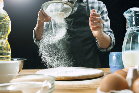 Professional male cook sprinkles dough with flour, prepares or bakes bread or pasta at kitchen tableの写真素材