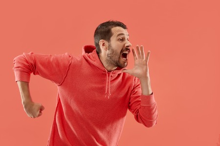 Do not miss. Young casual man shouting. Shout. Crying emotional man screaming on coral studio background. male half-length portrait. Human emotions, facial expression concept. Trendy colorsの写真素材