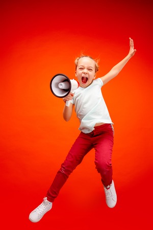 Beautiful young child teen girl jumping with megaphone isolated over redの写真素材