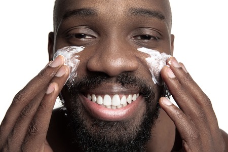 Young african-american guy applying face cream under his eyes on white background. Portrait of a young happy smiling african man at studio. High fashion male model.の写真素材